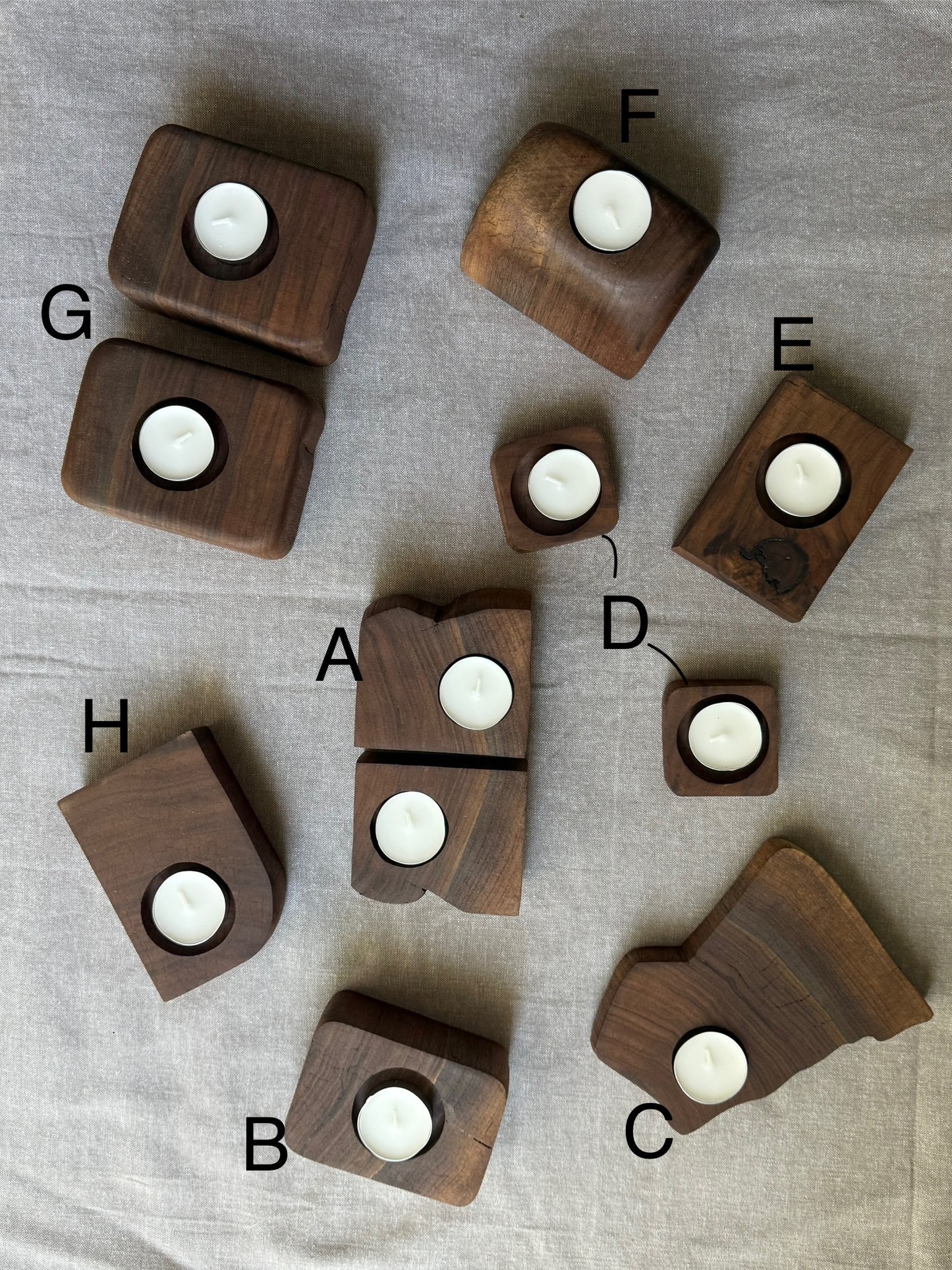 Eight lettered walnut candle holders arranged on linen backdrop – each with a white tea light candle, showcasing unique shapes