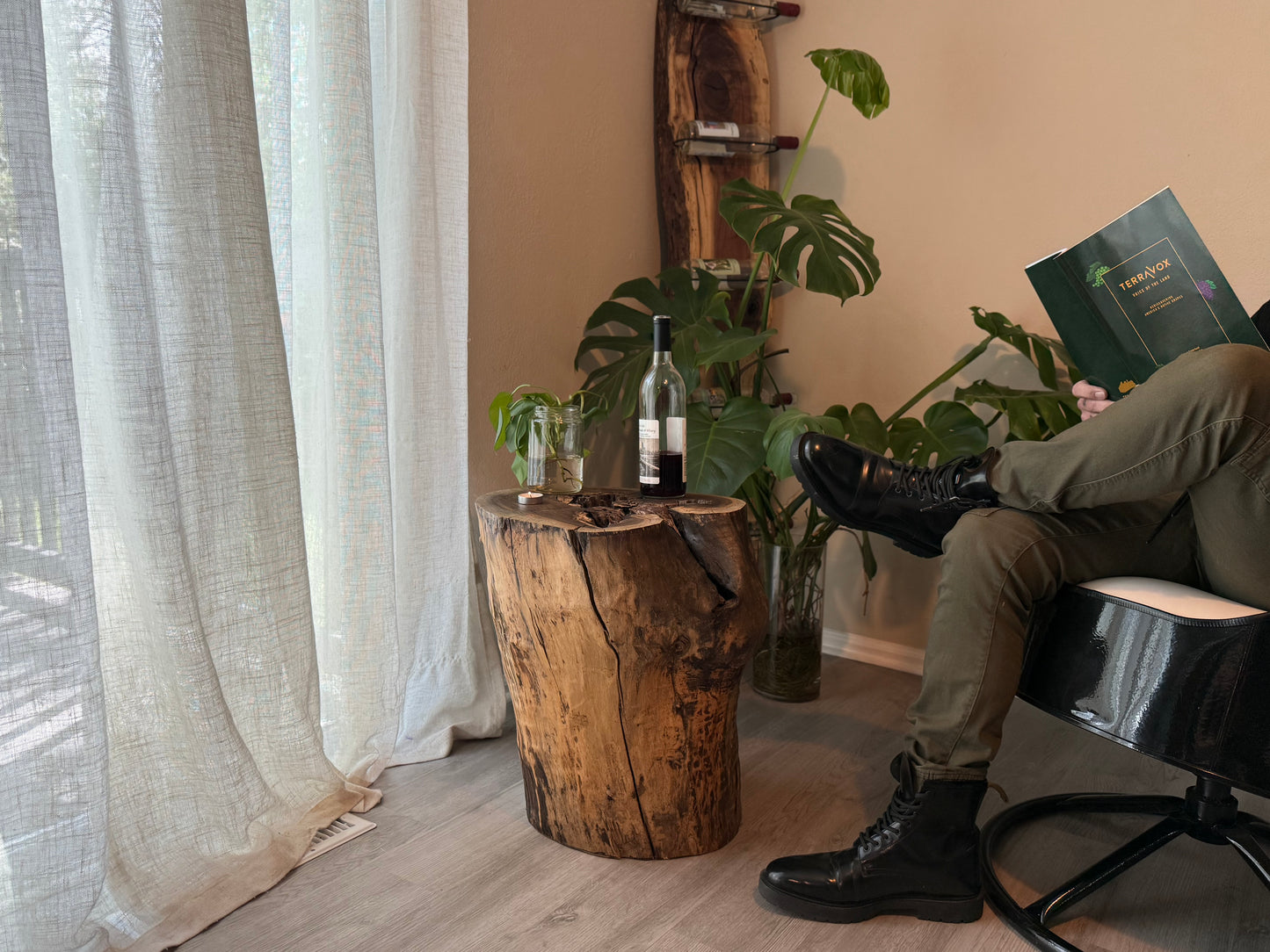 Person sitting on a chair reading a book in a room with plants and a natural wood stump table.